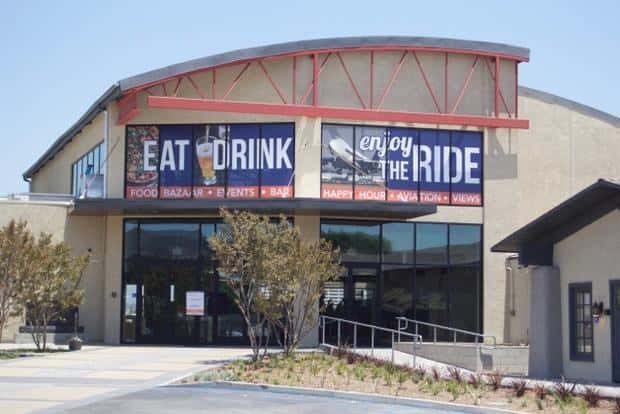 Exterior view of an aviation-themed restaurant and event venue with "Eat Drink Enjoy the Ride" signage and modern industrial architecture