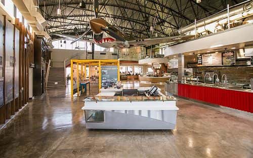Modern aviation-themed food hall interior with open kitchen counters, industrial design, and suspended vintage airplane display in a contemporary dining space