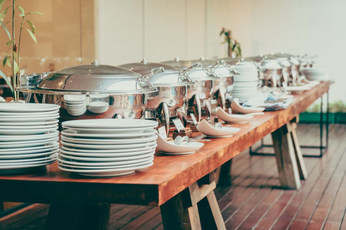 A long wooden table displays multiple silver chafing dishes and stacks of white plates, creating an inviting catering setup in a well-lit space.