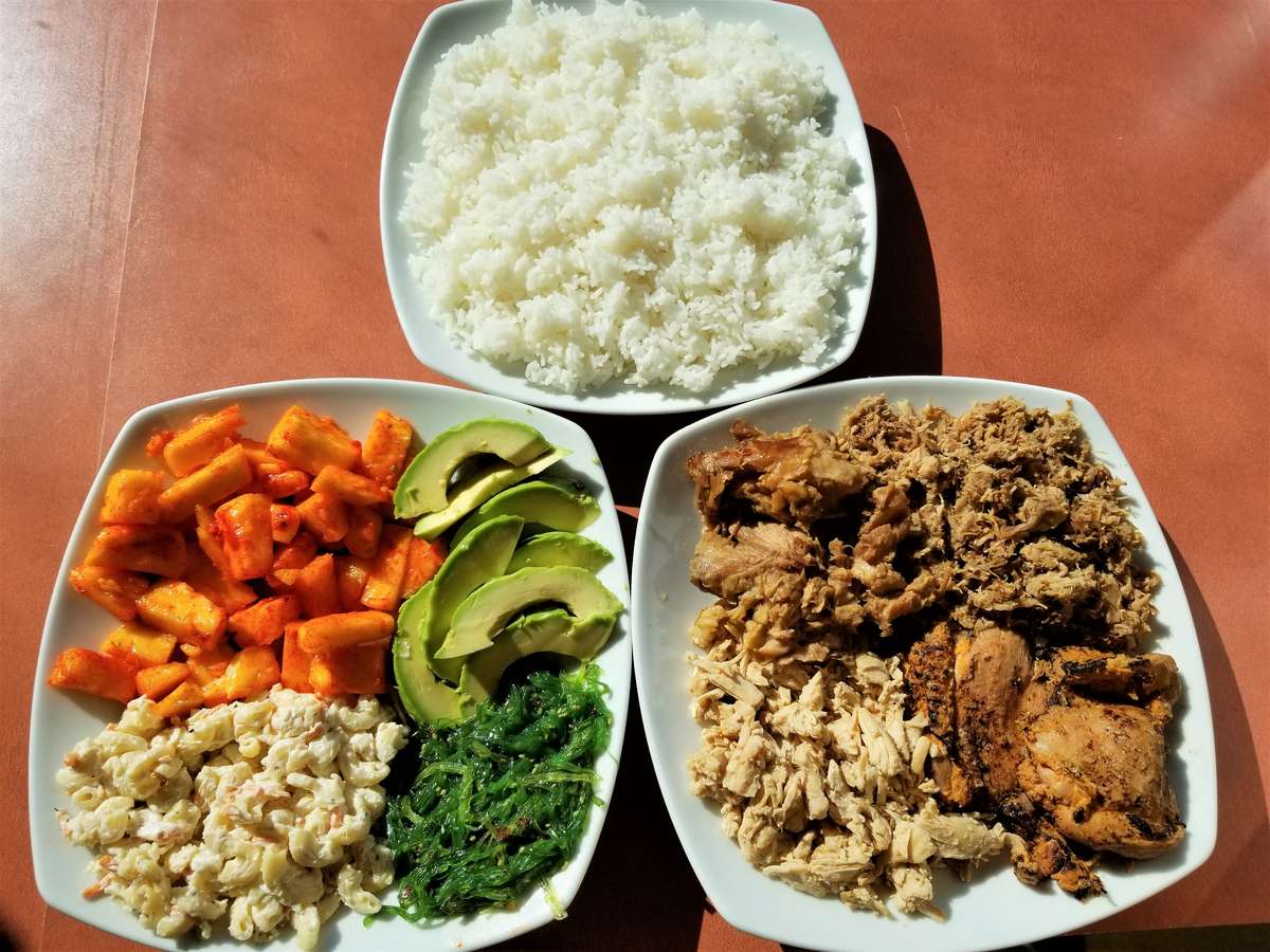 Three-plate Hawaiian-style meal featuring a bowl of steamed white rice, grilled meats including chicken and shredded pork, and a colorful side plate with avocado, macaroni salad, sweet potatoes, and seaweed salad.