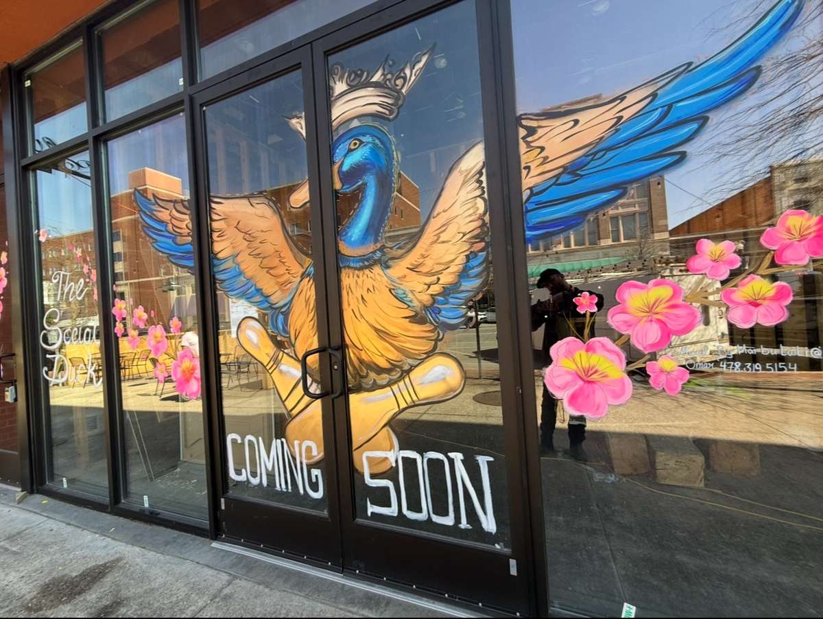 A painted sign on the window of The Social Duck announces 'Coming Soon' with a whimsical duck and bowling pin illustration.