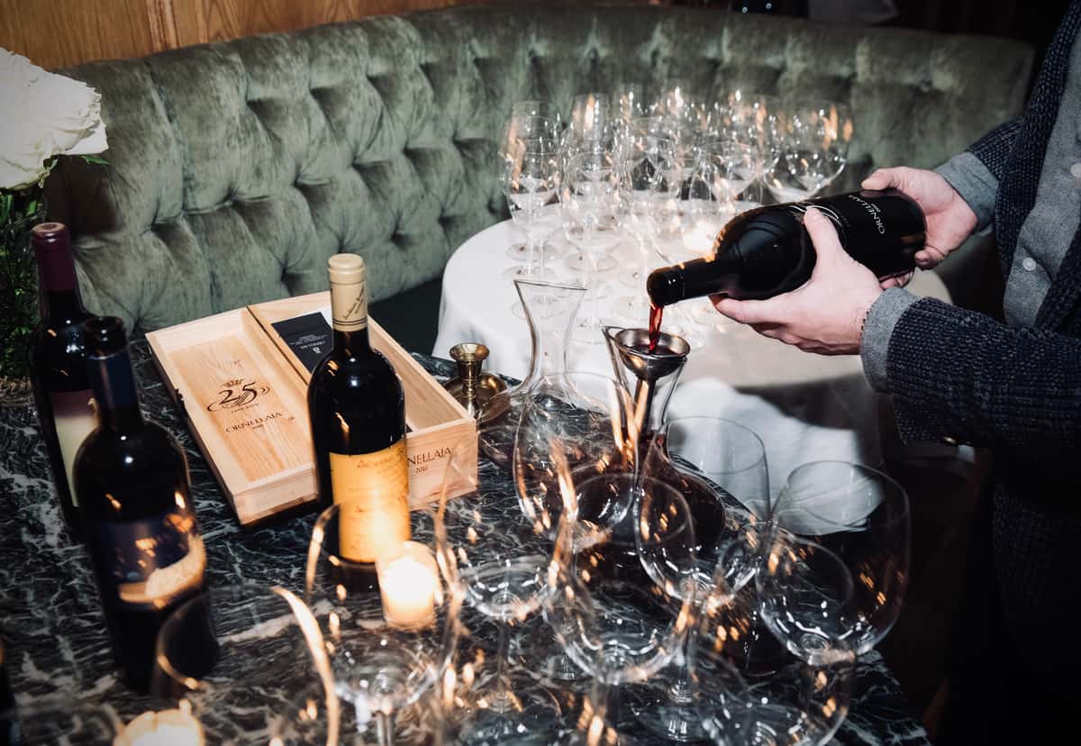 Sommelier pouring red wine into decanter beside bottles and empty glasses on marble table with candlelight