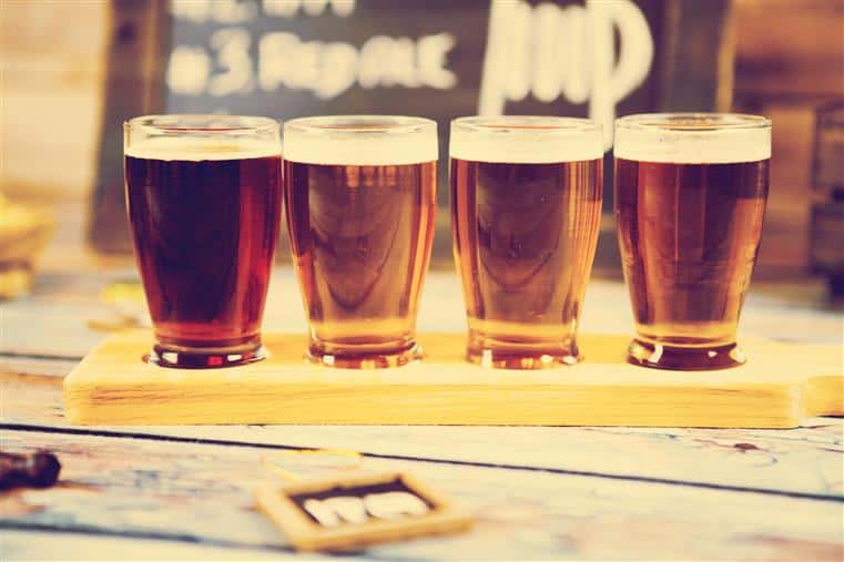Beer flight of four full beer glasses on wood table, chalkboard menu in the background.