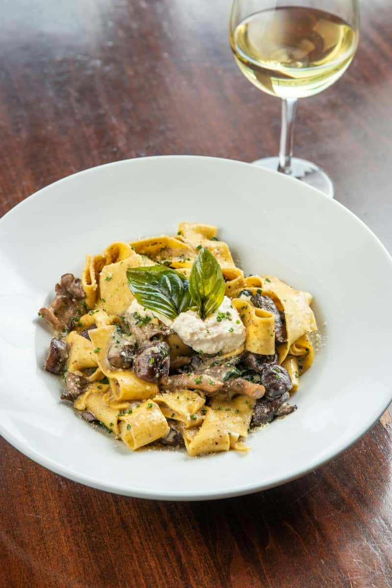 Pappardelle pasta with wild mushrooms, creamy ricotta, and fresh basil served with a glass of white wine in an upscale Italian restaurant