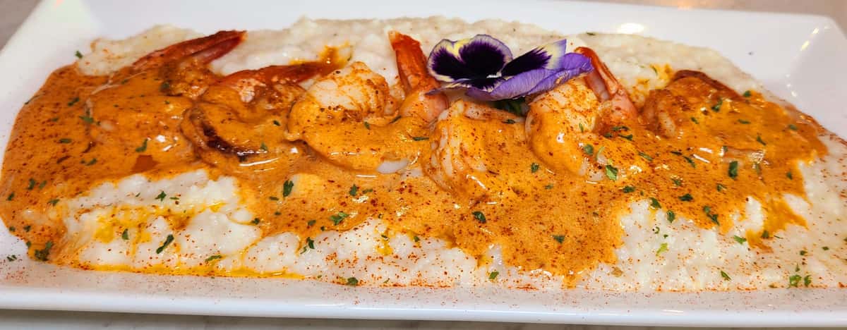 Creamy Southern-style grits topped with Cajun-spiced grilled shrimp in a rich, buttery sauce, finished with fresh herbs and a pansy garnish.