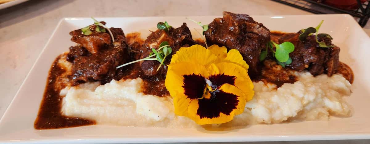 Tender braised oxtails in rich brown gravy over creamy mashed potatoes, garnished with microgreens and a vibrant edible flower.