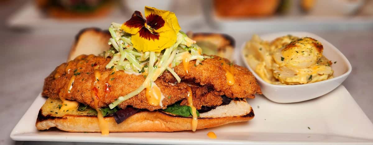 Crispy fried fish fillet on a toasted hoagie roll with slaw and a drizzle of sauce, served with a side of creamy potato salad and an edible flower.