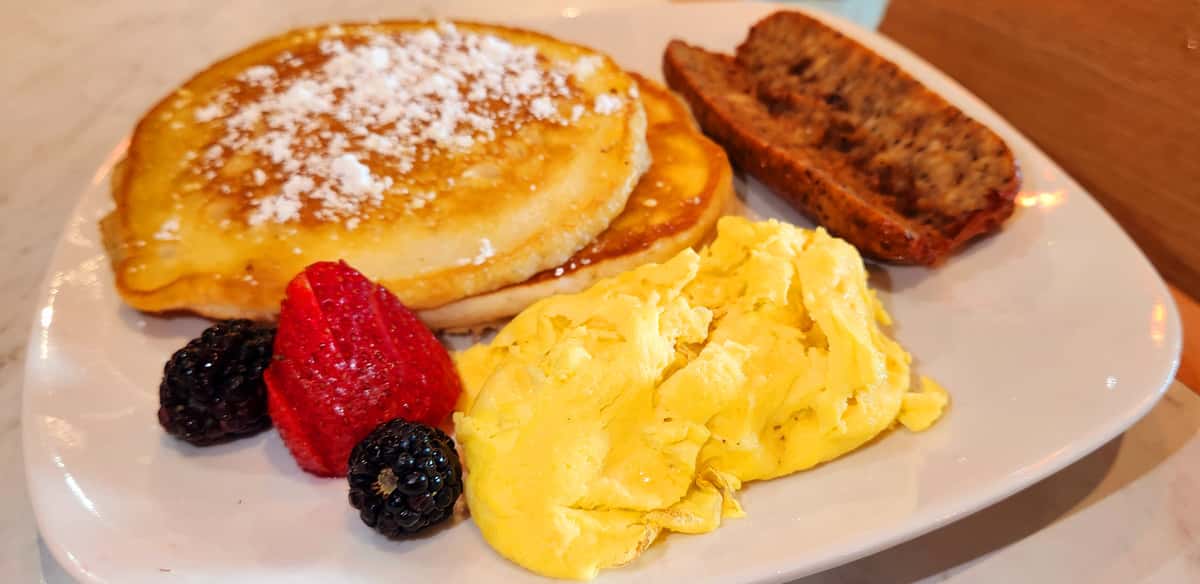 Fluffy pancakes topped with powdered sugar and syrup, served with scrambled eggs, fresh berries, and a slice of breakfast meatloaf.