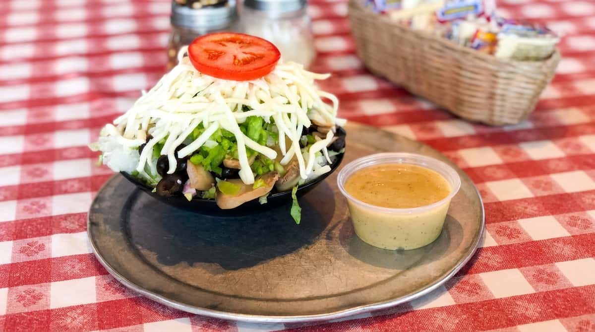 Taco salad in a black bowl, topped with shredded mozzarella, lettuce, and a tomato slice, with a side of dressing on a metal plate.