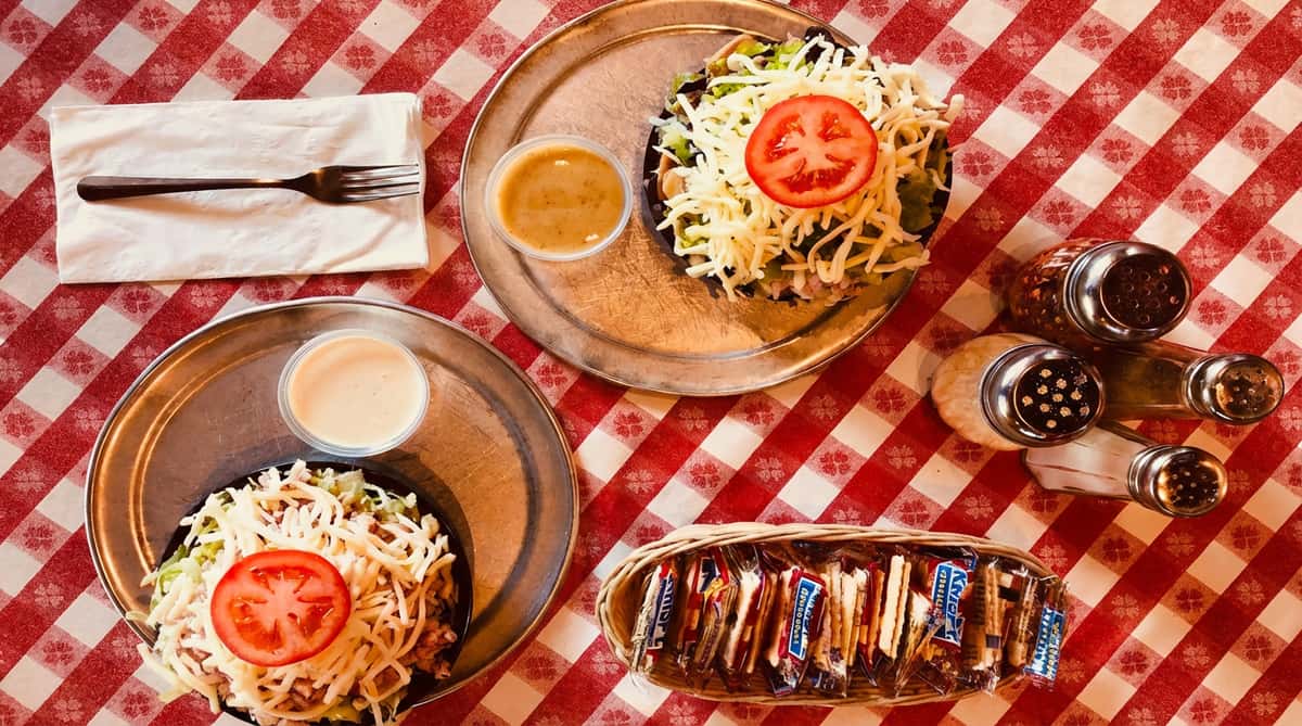 Two salads with lettuce, shredded cheese, and a tomato slice on metal plates with dressings, crackers, and shakers on a red and white checkered tablecloth.