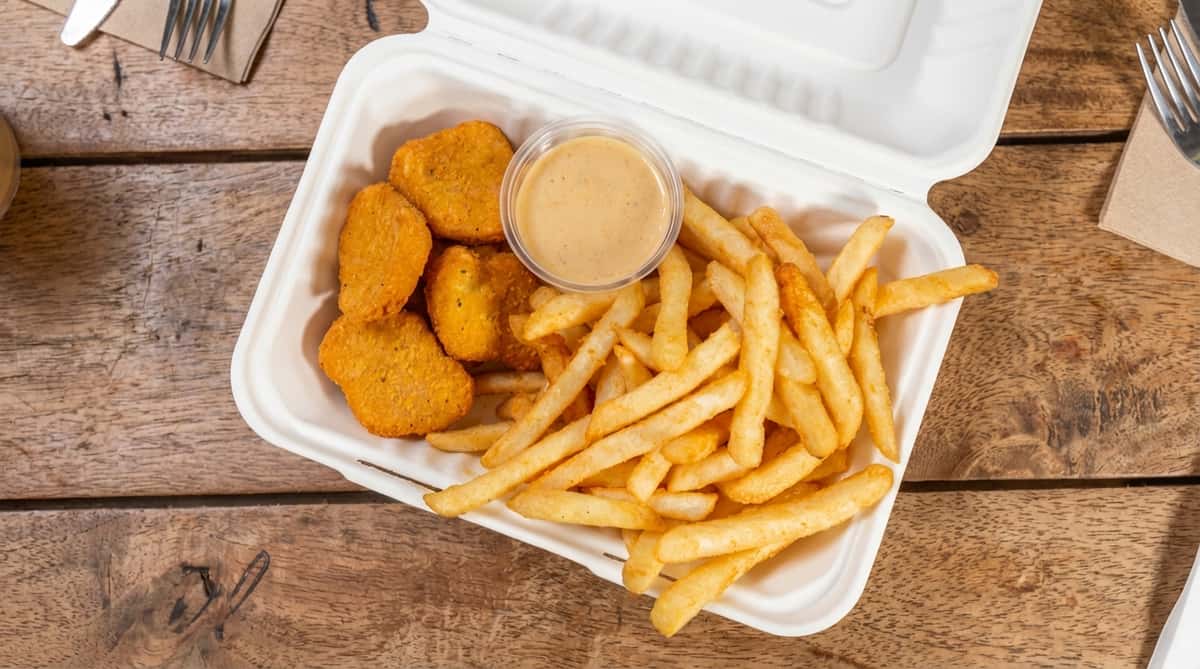 Chicken nuggets and french fries in a white to-go container with a cup of dipping sauce.