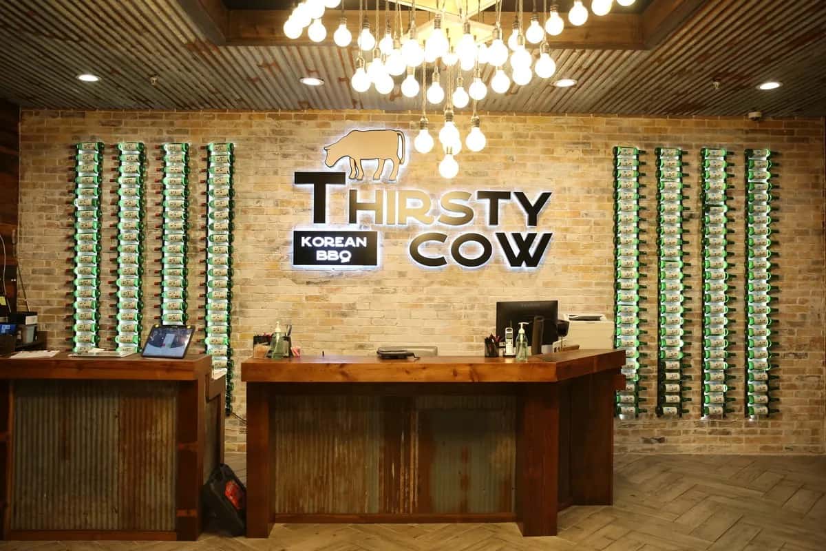 Thirsty Cow Korean BBQ front desk with illuminated logo, wooden counters, and walls lined with rows of soju bottles.