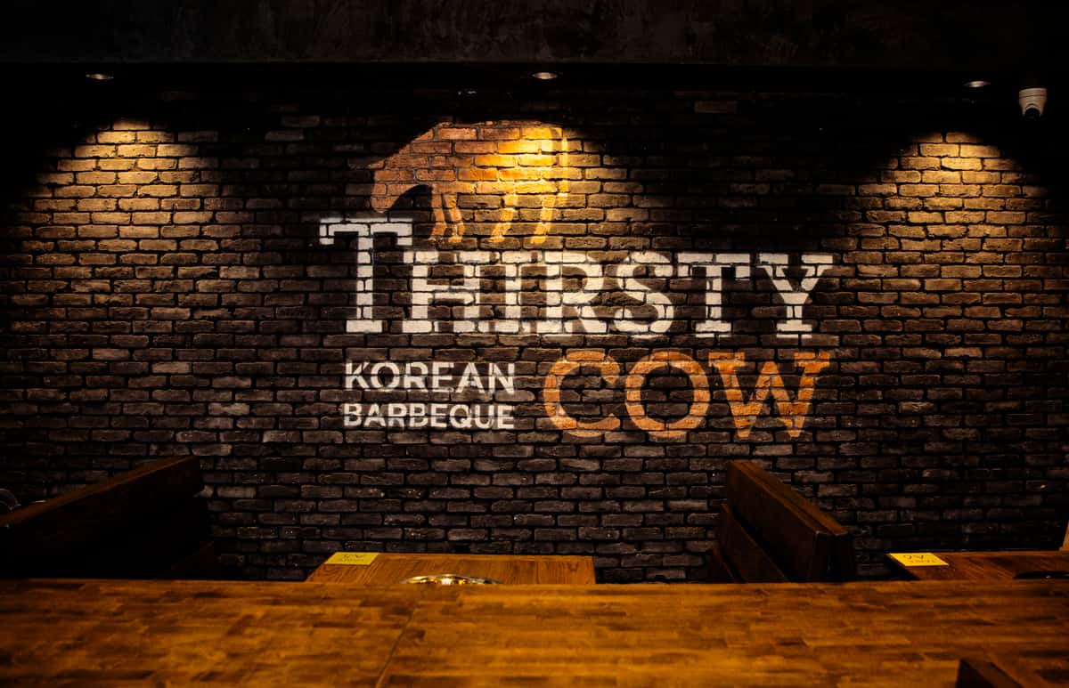 Thirsty Cow Korean Barbeque logo painted on a brick wall, lit by warm spotlights above wooden dining booths.
