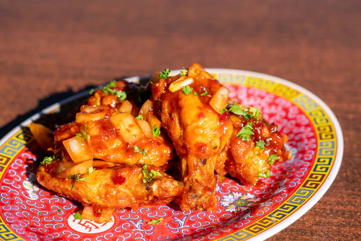 A plate of crispy chicken wings coated in a glossy sweet and spicy sauce, garnished with chopped green onions and sesame seeds. The wings are served on a vibrant decorative platter, creating a bold and flavorful presentation with a savory, sticky finish.