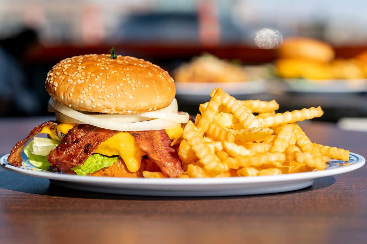 A classic cheeseburger served on a sesame seed bun with melted cheese, fresh toppings, and a juicy beef patty, paired with a generous side of crispy golden French fries. The warm lighting and close-up presentation give the meal a rich, satisfying diner-style appeal.