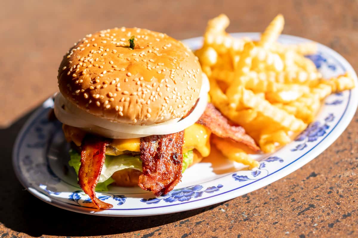 A gourmet burger served on a toasted sesame seed bun, layered with crispy fried chicken, melted cheese, fresh lettuce, and sauce. The burger is plated alongside a generous portion of golden French fries on a patterned dish. The warm lighting and close-up angle highlight the crispy textures and rich flavors, giving the image an appetizing classic diner-style feel.