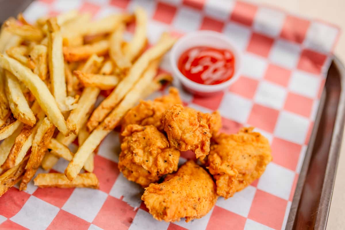 A child's portion of crispy chicken nuggets served with fries and ketchup on a checkered tray at The Butcher Shop restaurant
