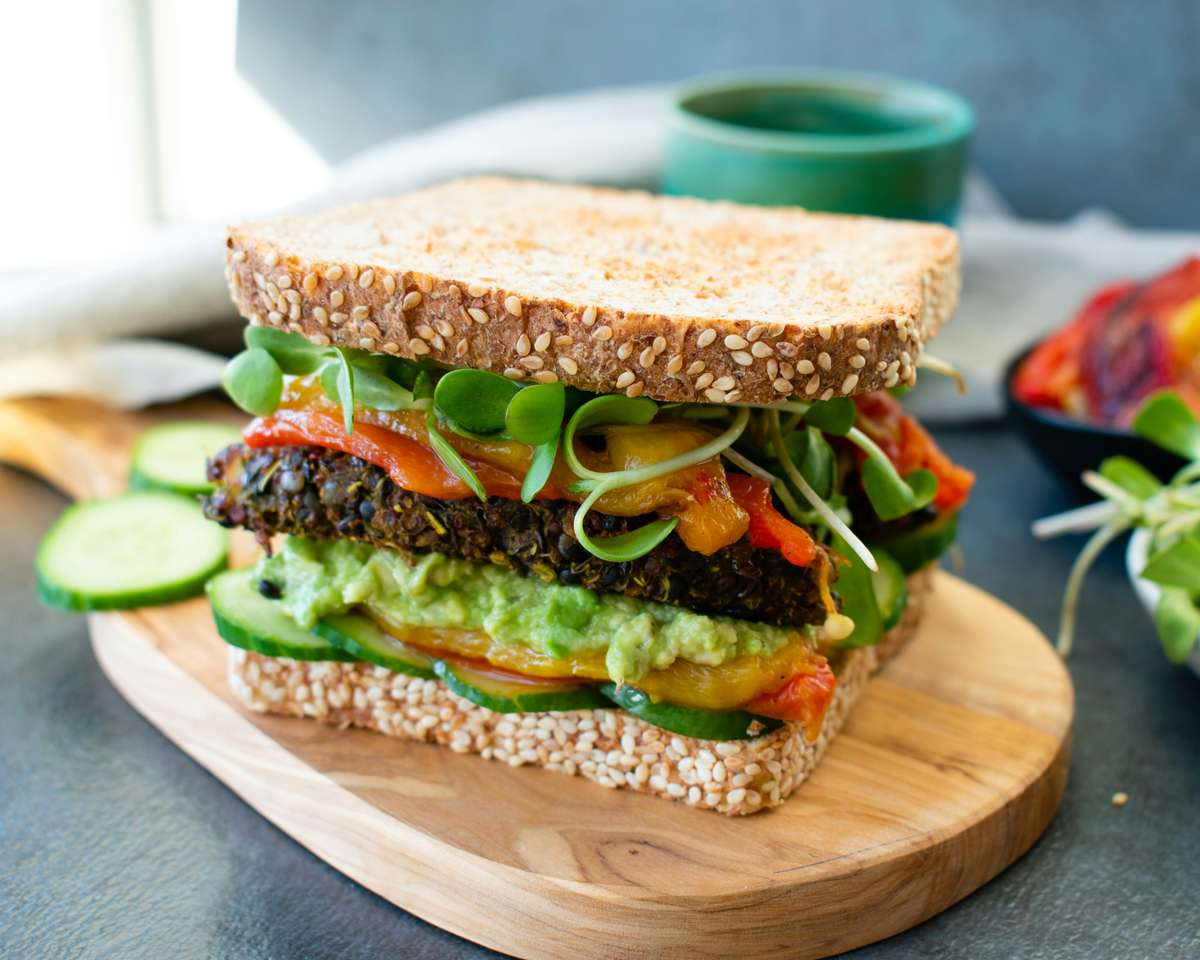 Vegan sandwich with seed-crusted whole grain bread, black bean patty, avocado spread, cucumber, roasted peppers, and microgreens.