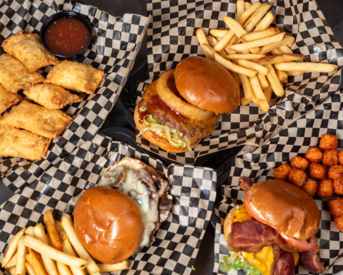 Overhead view of burgers, fries, sweet potato tots, fried wontons, and dipping sauce arranged on serving trays.