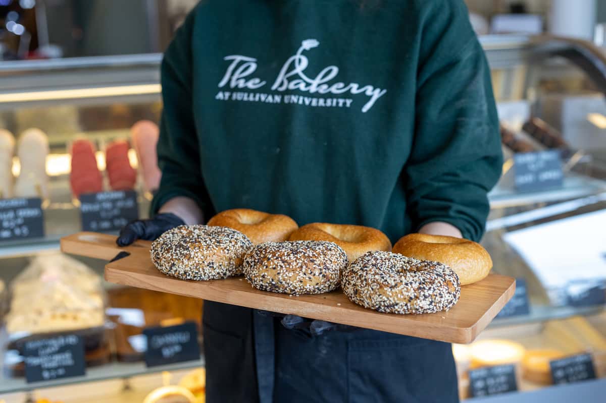 A person holds a wooden tray of bagels, three with everything-seasoning. The person wears an apron and a sweatshirt that reads: "The Bakery at Sullivan University".