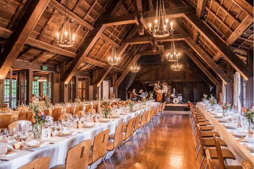 Rustic luxury banquet hall with long communal tables, exposed wood beams, and warm chandelier lighting
