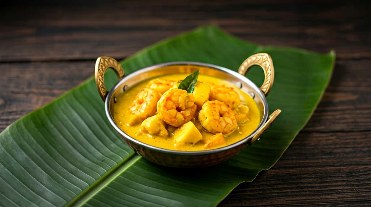 Shrimp curry with mango and a green leaf garnish in a brass bowl on a banana leaf.