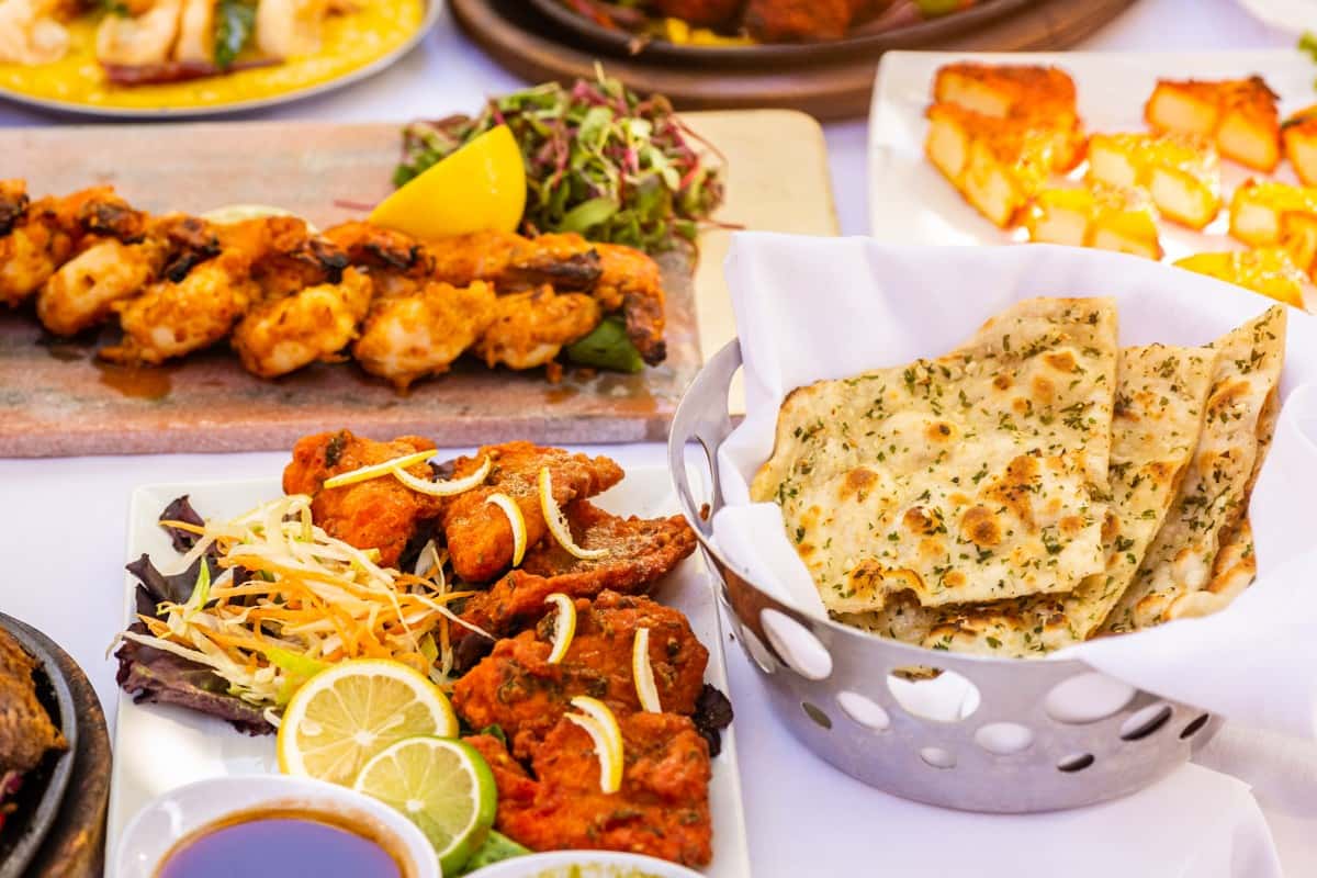 Basket of garlic naan served with tandoori shrimp skewers, fish pakora, and a side of sliced lemon and salad.