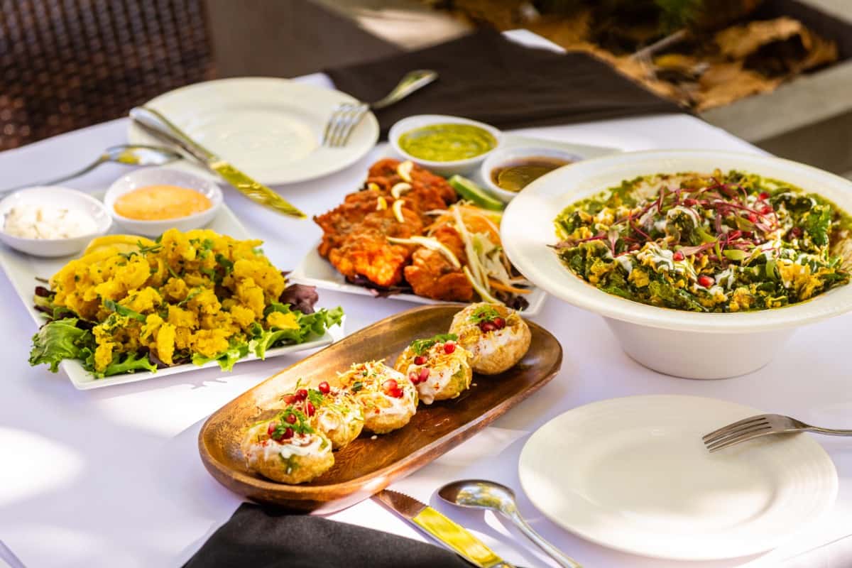 Indian lunch table with palak chaat, dahi puri, tandoori chicken tikka, crispy okra, chutneys, and yogurt dips on white linens