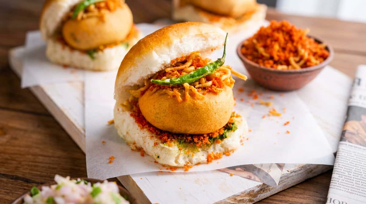 Vada Pav, a deep-fried potato fritter in a bun with green chutney, spicy powder, crispy noodles, and a green chili on top.