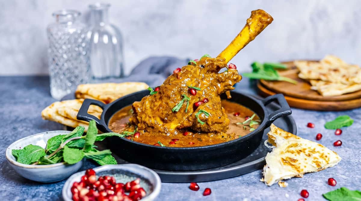 Lamb shank curry with pomegranate seeds and mint in a black cast iron dish, served with flatbread.