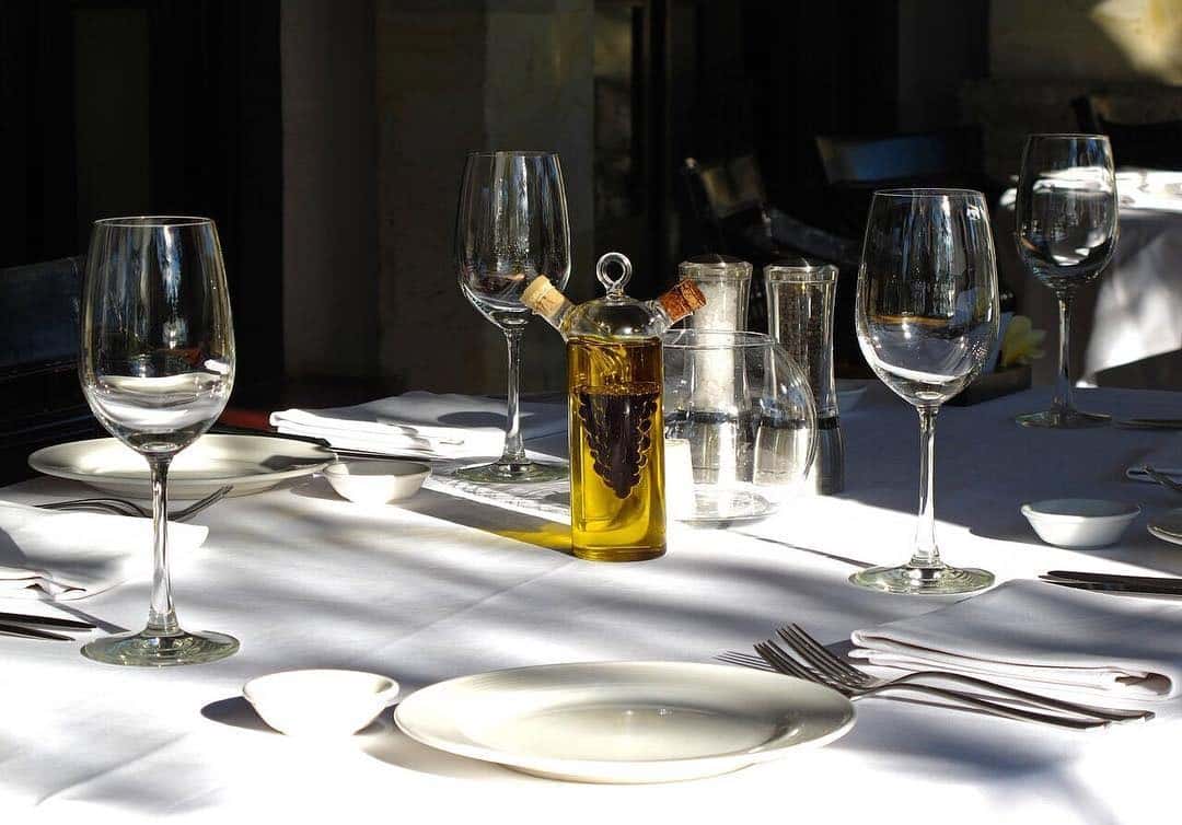 Elegant outdoor dining table set with wine glasses, plates, cutlery, and a decorative oil and vinegar cruet in the sunlight.