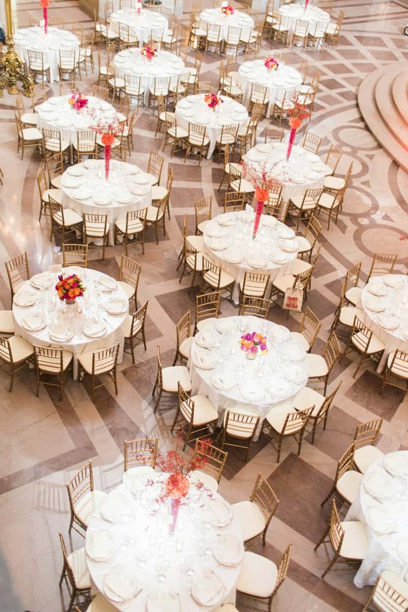 Elegant banquet hall with round tables set for an event, decorated with white linens, gold chairs, and vibrant floral centerpieces.