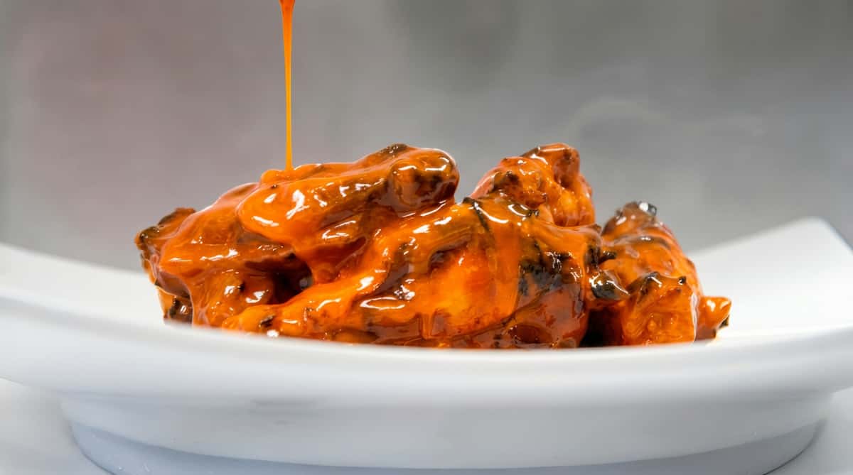 Chicken wings covered in orange buffalo sauce with an additional stream of sauce pouring over them, on a white plate.