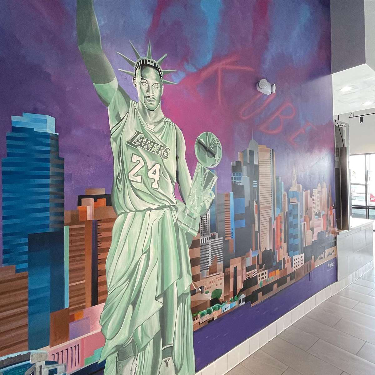 Colorful mural of basketball player in a Lakers 24 jersey styled as the Statue of Liberty holding a championship trophy over a city skyline.