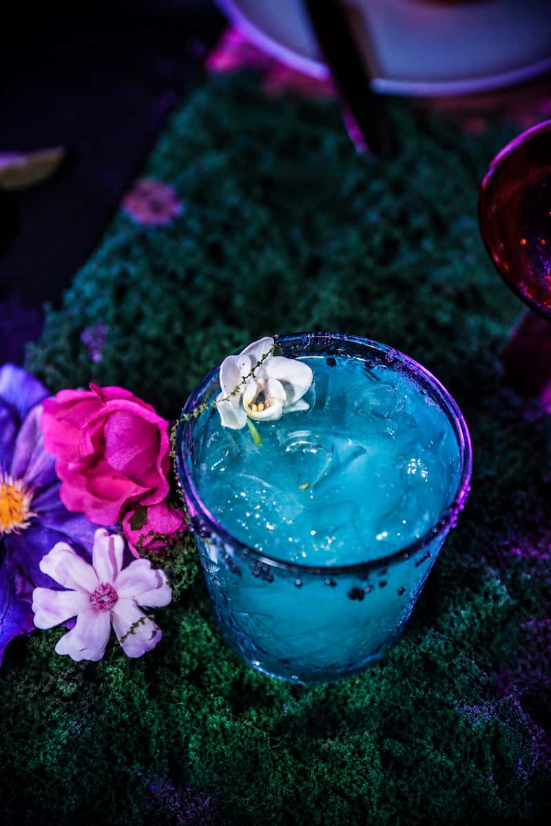 Turquoise Mesa cocktail beside pink and purple flowers under blacklight-style lighting