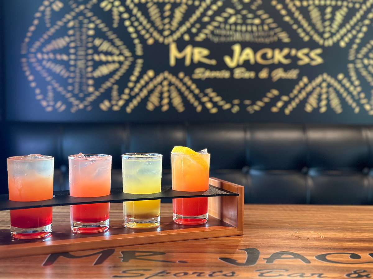 Colorful cocktails sit in a wooden holder in front of a black wall with gold designs. The backdrop reads "Mr. Jack's Sports Bar & Grill."