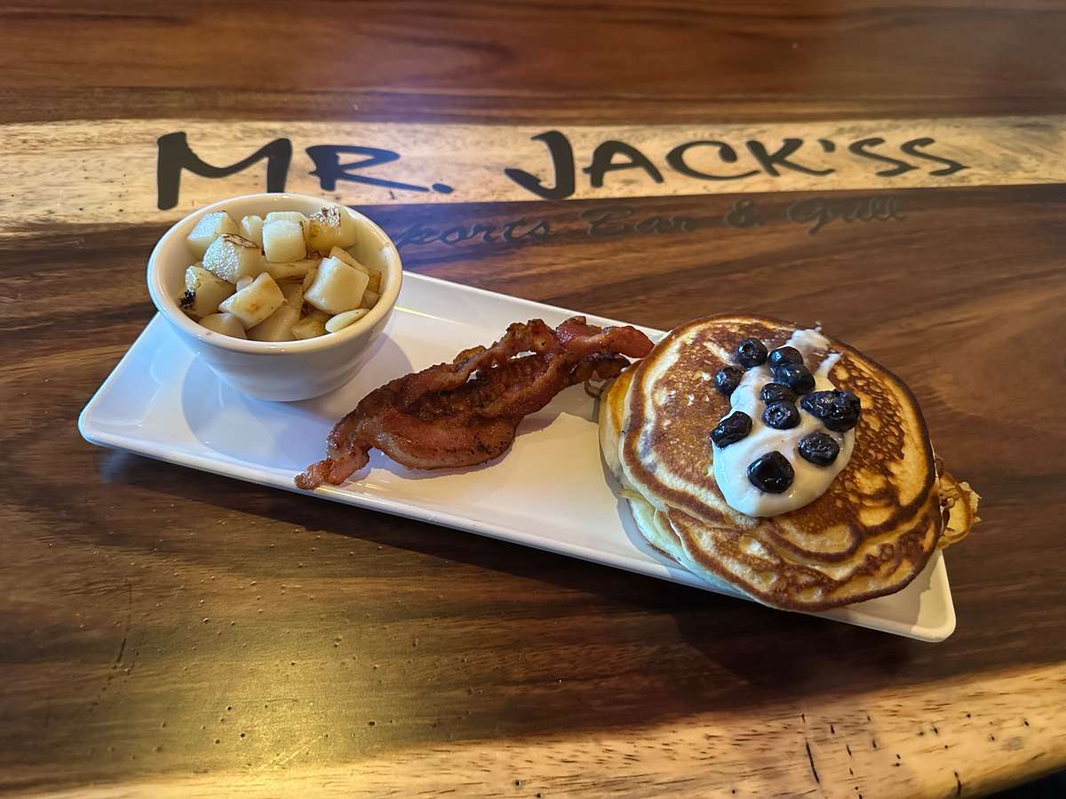 Here's a tasty breakfast from Mr. Jack'ss Sports Bar & Grill. A stack of pancakes with berries and cream, bacon, and fried potatoes are served on a white plate.