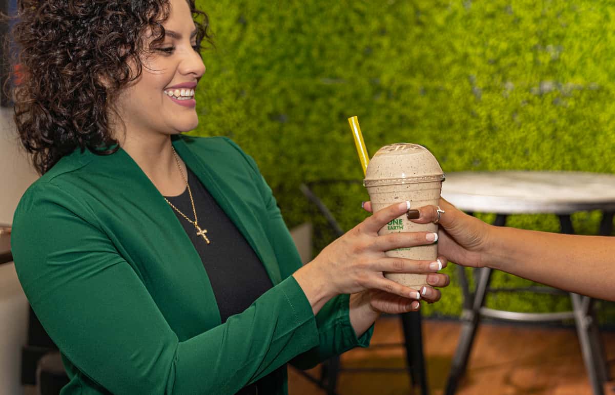 Smiling woman in a green blazer receives a smoothie with a yellow straw, handed to her against a vibrant green background.