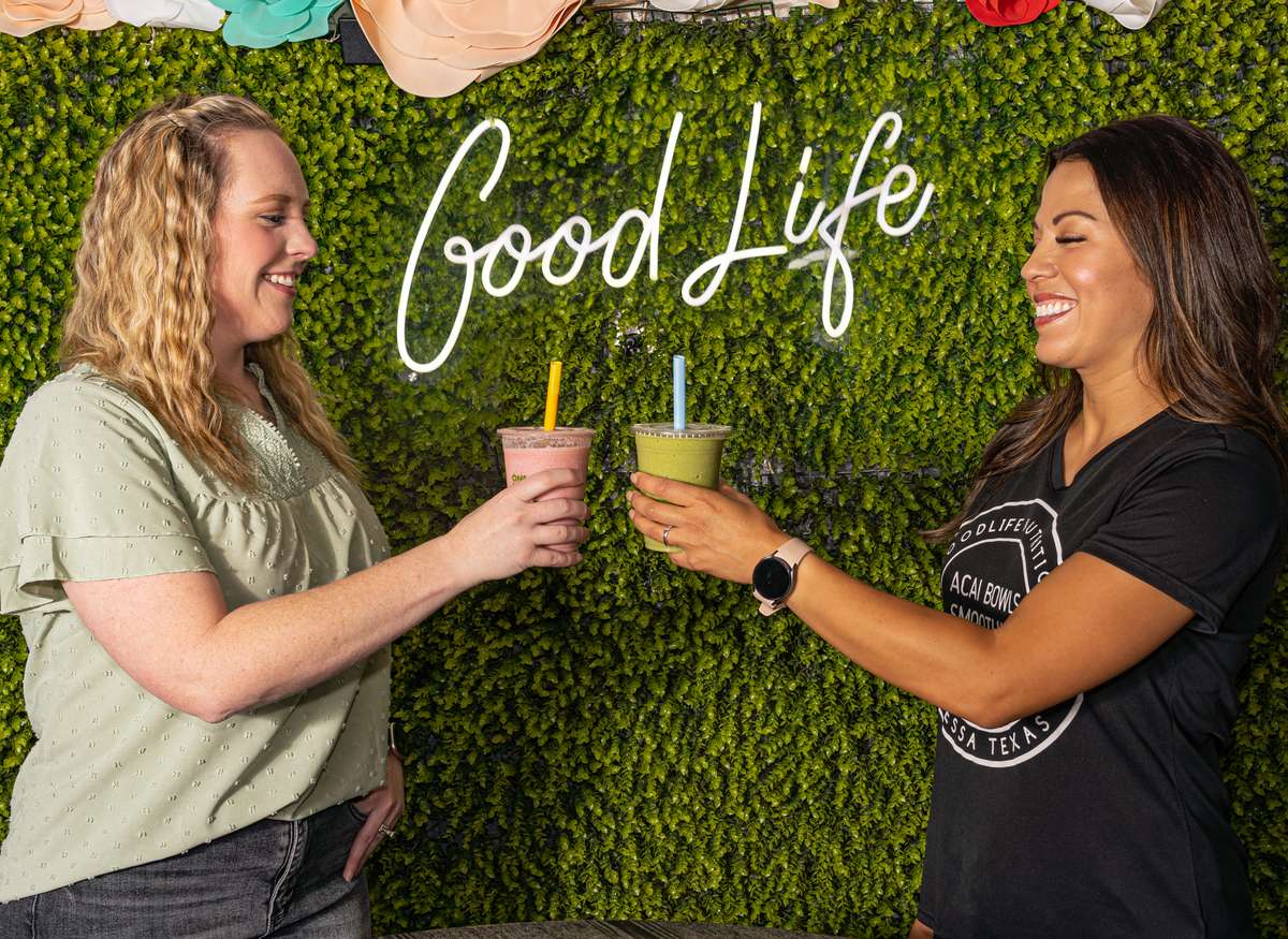 Two smiling women clink smoothies in front of a green wall with a "Good Life" neon sign and colorful paper flowers above.