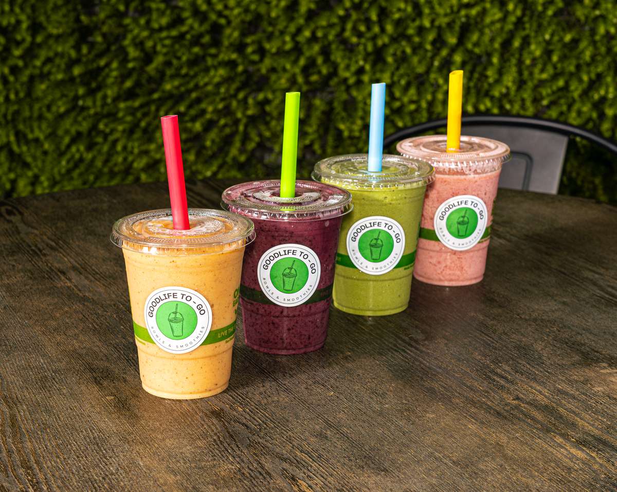 Four colorful Good Life To Go smoothies with vibrant straws on a rustic wooden table against a green plant wall.