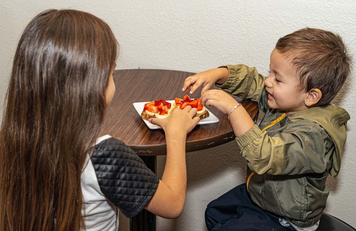Two young children sitting at a small table, happily sharing toast topped with banana slices and fresh strawberries.
