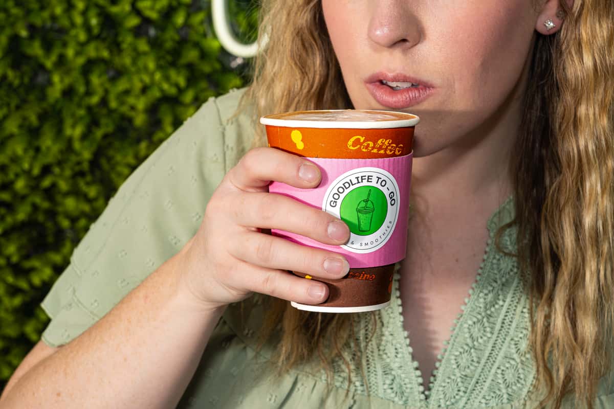 Woman sipping a hot drink from a GoodLife To-Go cup with a pink sleeve and latte art on top.