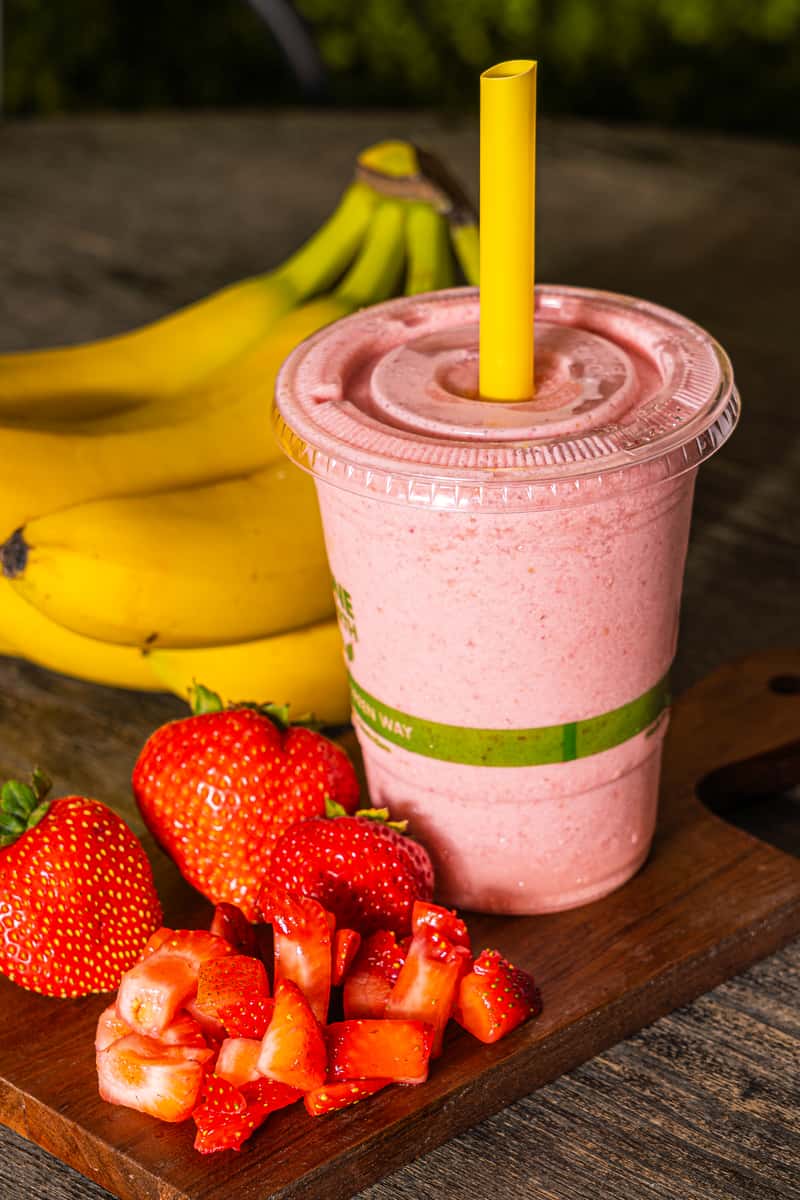 Strawberry banana smoothie in a plastic cup with fresh strawberries and bananas on a wooden surface.
