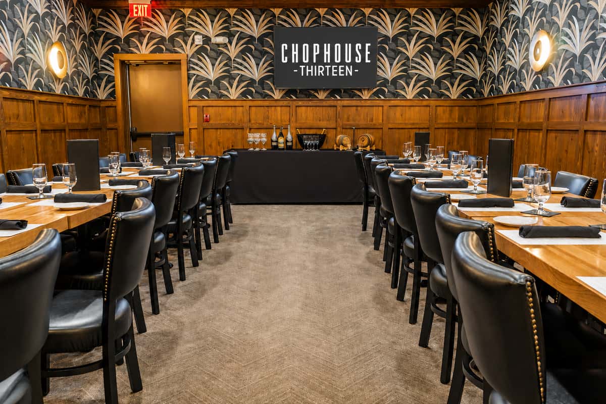 **Alt text:** Private dining room at Chophouse Thirteen featuring long wood tables with black leather chairs, elegant place settings, warm wood paneling, art deco wallpaper, a beverage service station, and a television displaying the Chophouse Thirteen logo, arranged for a private event or corporate gathering.