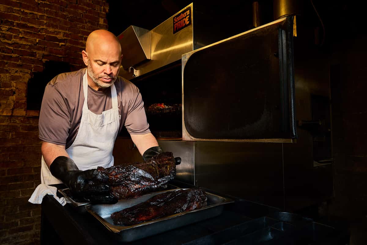 A pitmaster carefully tends to a tray of freshly smoked barbecue meats inside a commercial smoker. Surrounded by the warm glow of the kitchen, the scene highlights the craftsmanship and attention to detail that go into preparing authentic, slow-cooked barbecue with rich flavor and tender texture.
