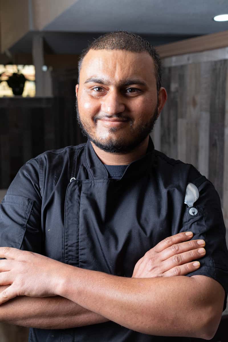 A chef stands smiling confidently with arms crossed, wearing a black chef coat with a fork emblem on the sleeve.