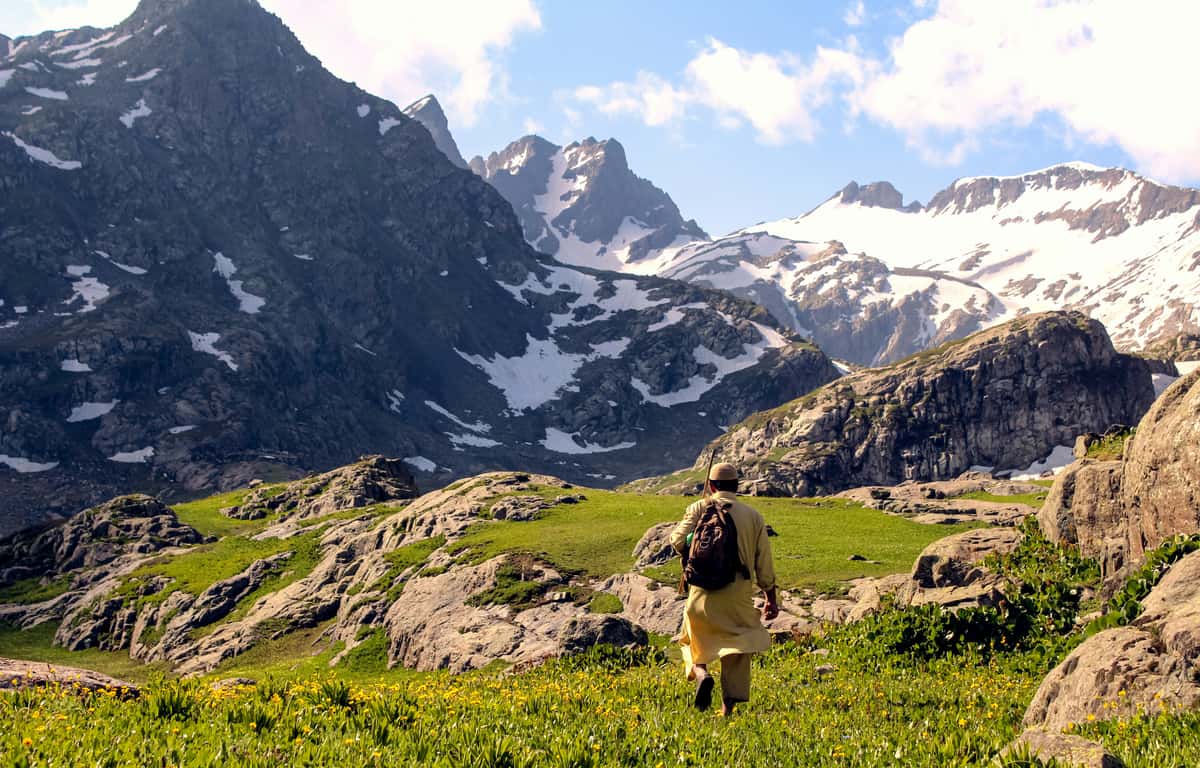 A person in traditional attire with a backpack hikes through a lush alpine meadow surrounded by rocky terrain and snow-capped mountains under a bright sky.