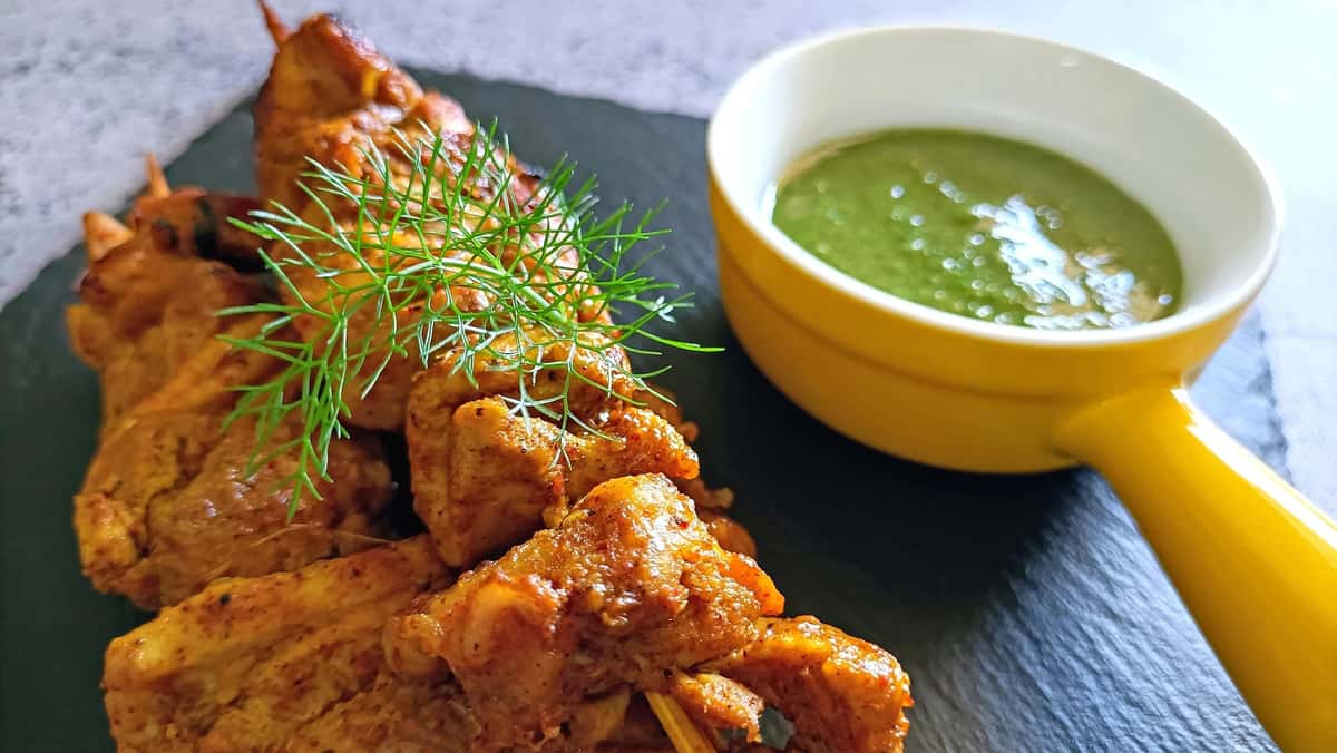 This image shows skewered grilled chicken tikka garnished with fresh herbs, served alongside a small bowl of green chutney in a yellow-handled dish. The presentation is elegant and modern, with a slate serving board enhancing the dish's vibrant colors.