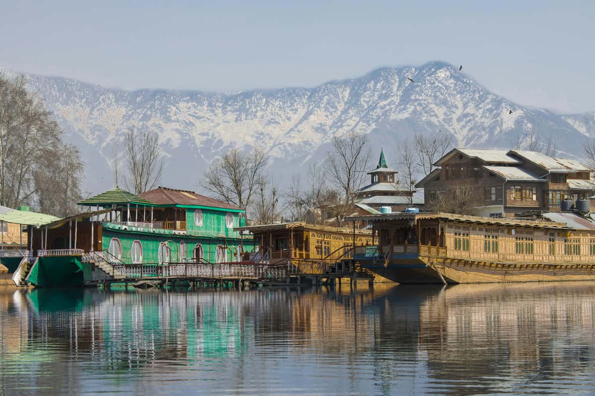This image showcases traditional wooden houseboats on a serene lake, likely Dal Lake in Srinagar, Kashmir. The backdrop features snow-capped Himalayan mountains under a clear blue sky, with leafless trees hinting at a winter or early spring setting. The calm water beautifully reflects the vibrant colors and detailed architecture of the houseboats.