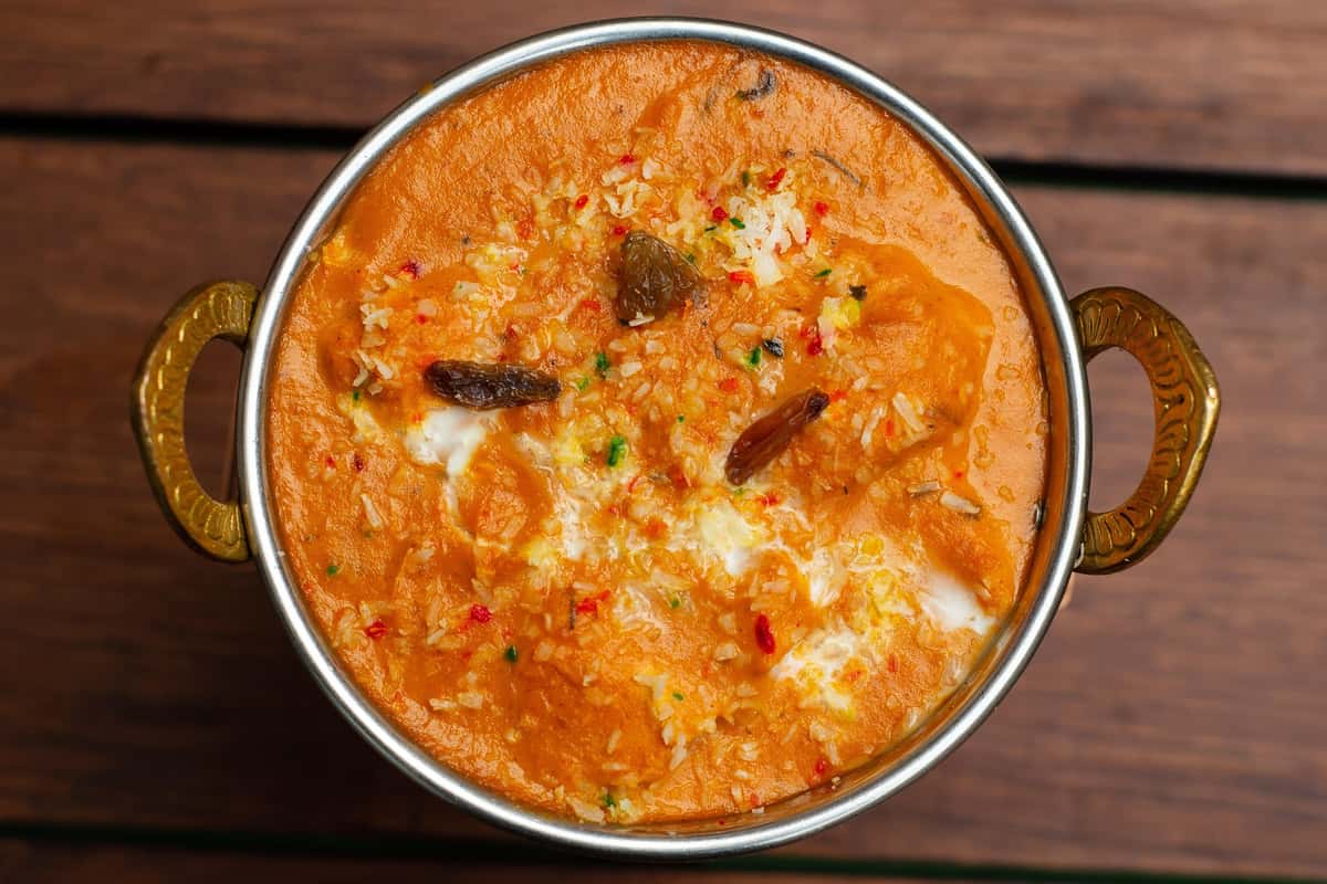 Rich and creamy Indian curry topped with grated paneer, raisins, and a drizzle of cream, served in a traditional brass-handled bowl.